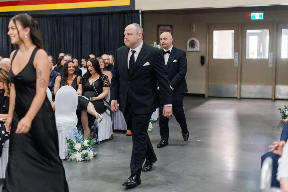 Men in black suits walk down the aisle at an elegant wedding ceremony in a decorated banquet hall.