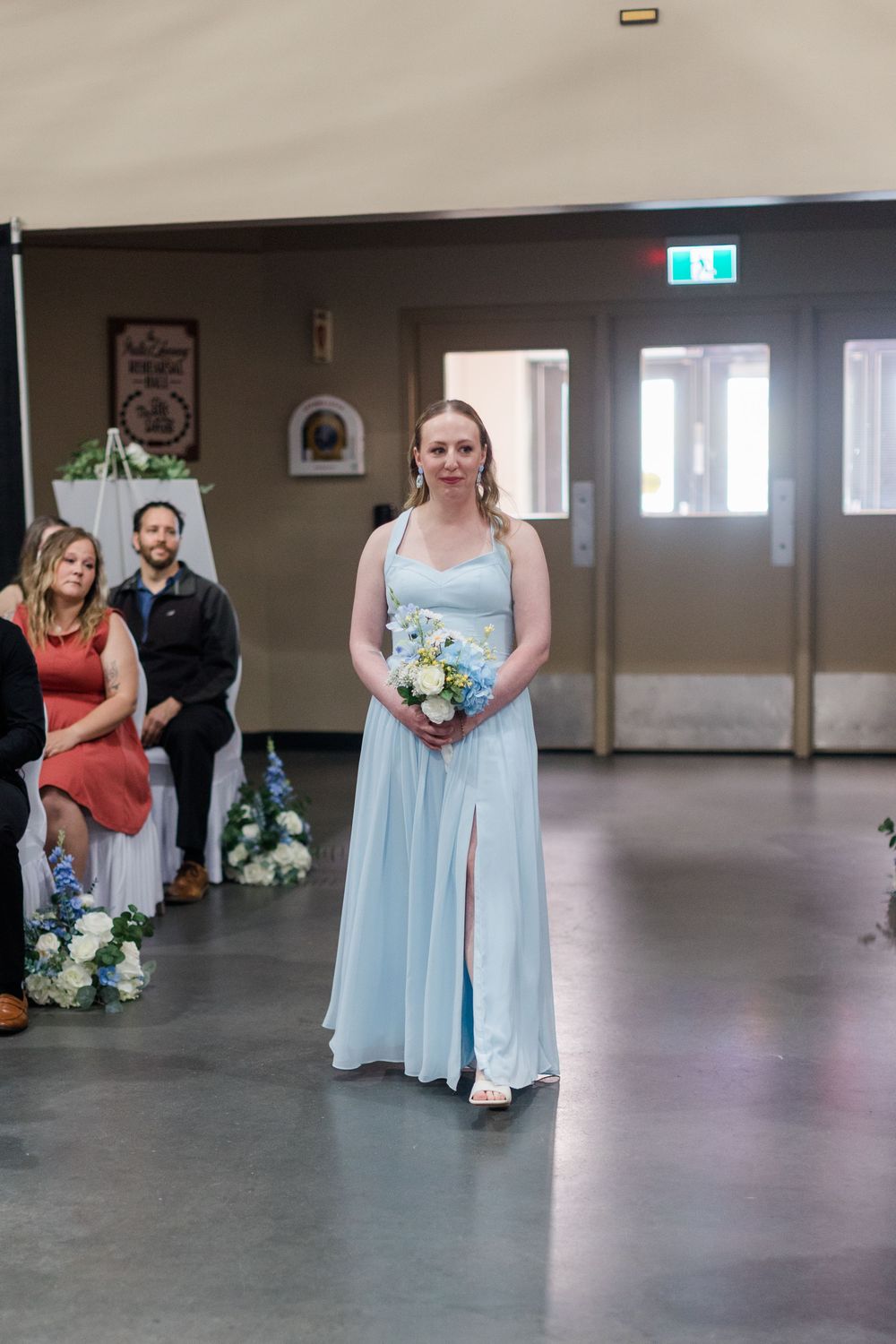 Bridesmaid in light blue gown walks down the aisle holding a bouquet at an indoor wedding ceremony.