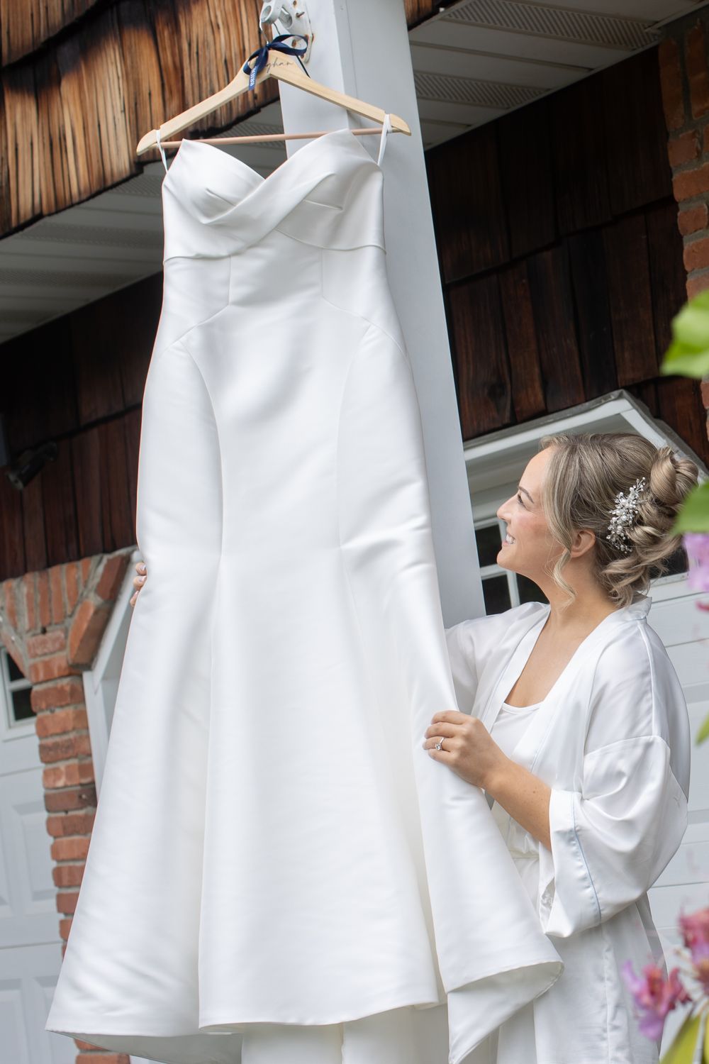 Bride in white robe admiring her strapless wedding gown hanging outside on a wooden hanger.