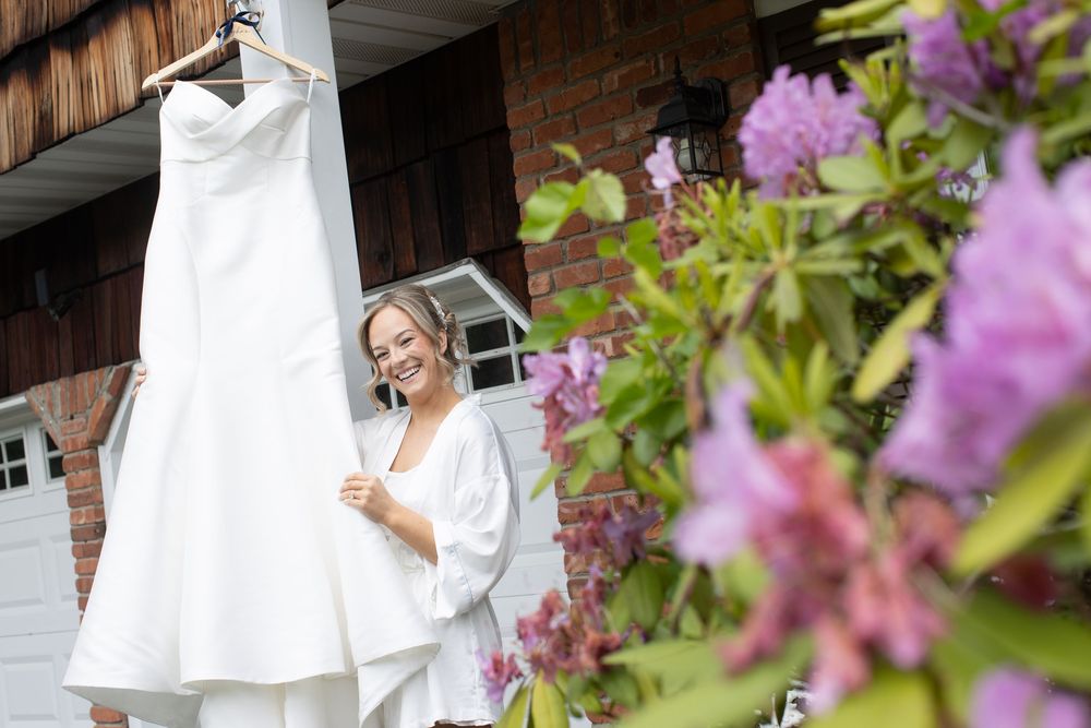 Smiling bride in white robe admiring her wedding dress hanging outside a home with blooming purple flowers.