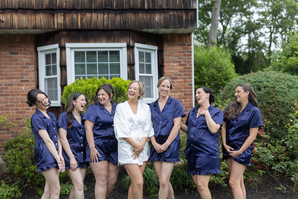 Bride and six bridesmaids laugh together outdoors in matching navy pajama sets before the wedding ceremony.