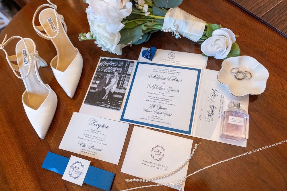 Wedding flat lay featuring ivory heels, floral bouquet, blue and white invitations, rings, and perfume on wooden table.