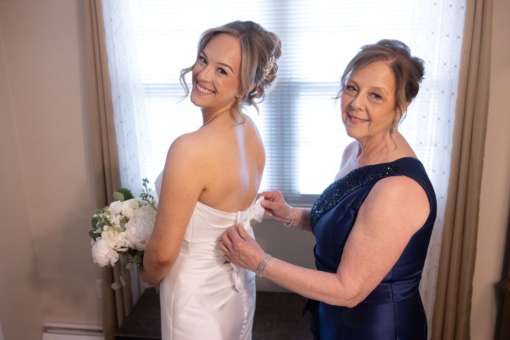 Bride in white gown smiles as older woman in navy dress buttons up the back before the wedding ceremony.