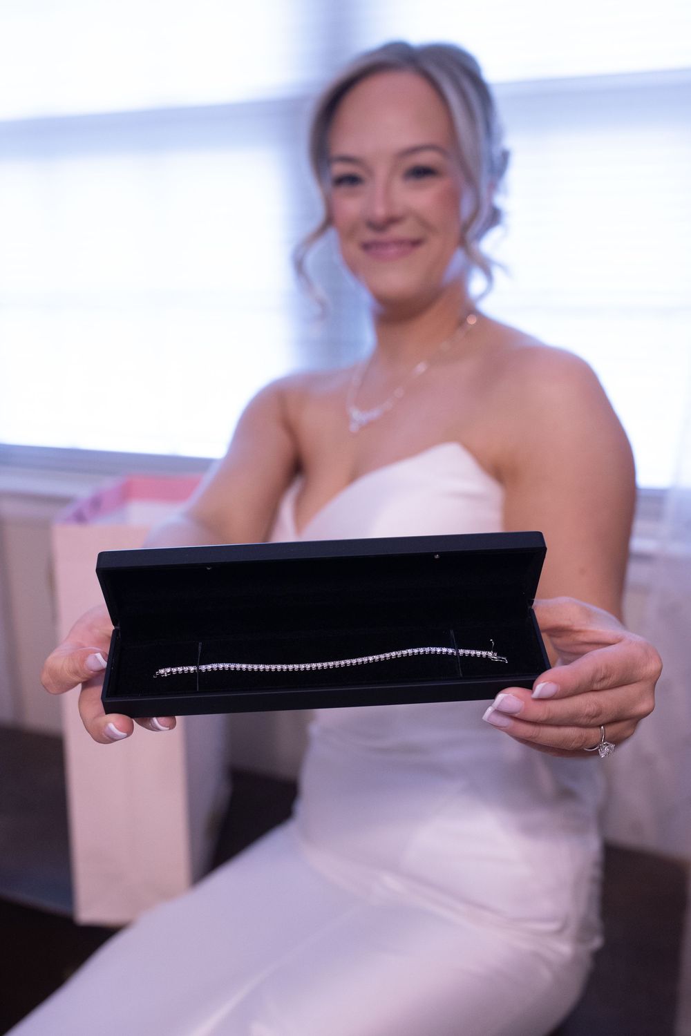 Smiling bride in white gown displaying a diamond tennis bracelet in a black jewelry box.