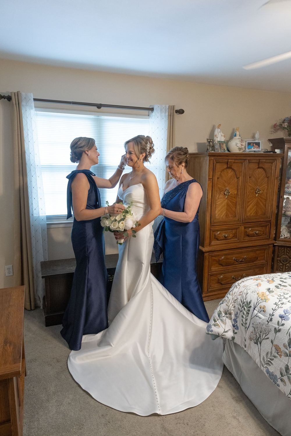 Bride in white gown holds bouquet as two bridesmaids in navy blue dresses help her prepare in a cozy bedroom.