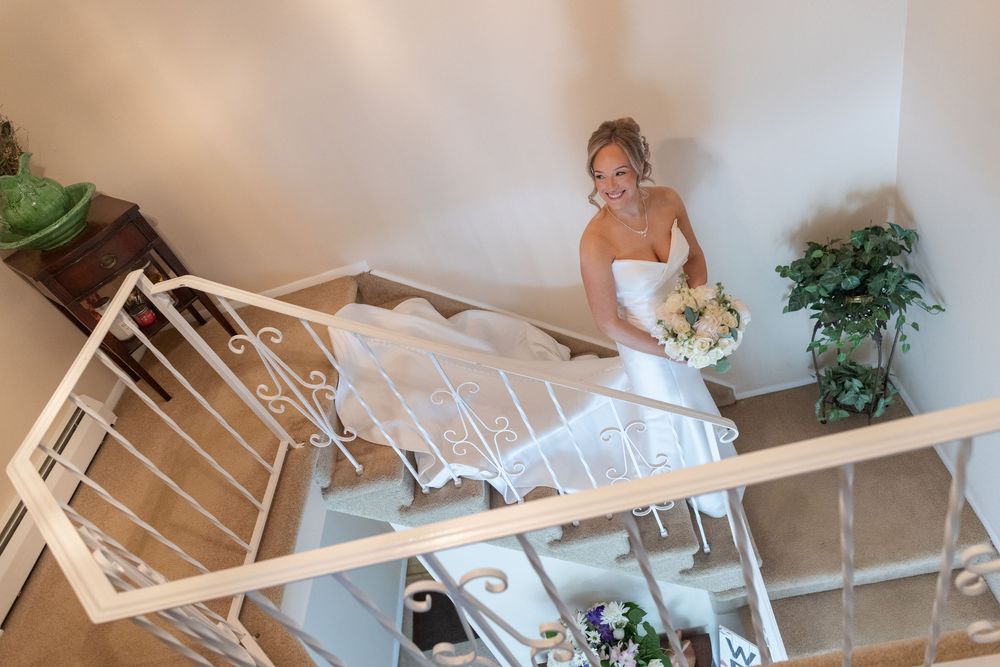 Elegant bride in strapless wedding gown descending a white staircase holding a bouquet.