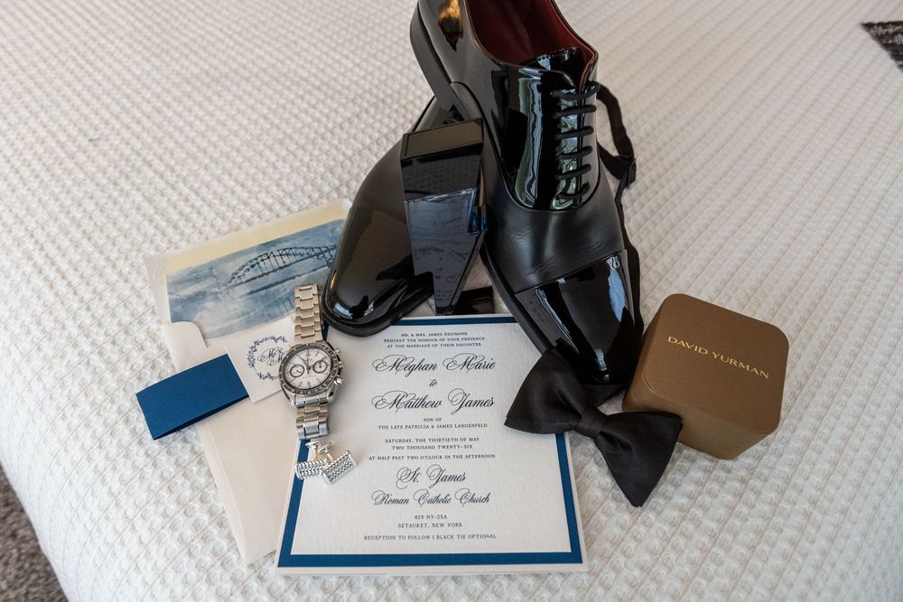 Groom wedding details flat lay featuring black patent shoes, bow tie, watch, and formal invitation.
