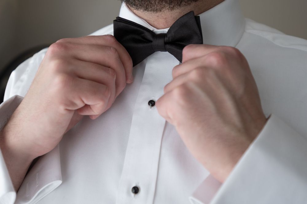 Groom adjusts his black bow tie on a white tuxedo shirt before the wedding ceremony.
