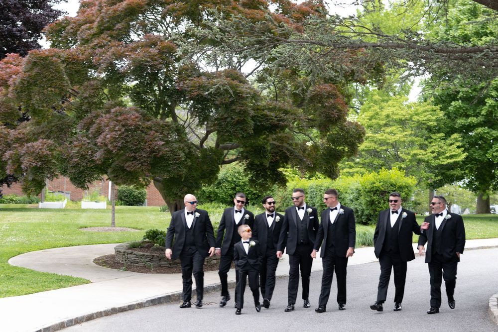 Groom and groomsmen in black tuxedos with sunglasses walk confidently along a garden pathway together.