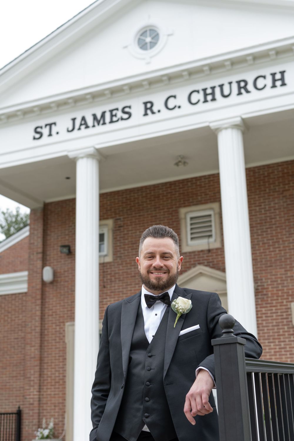 Smiling groom in black tuxedo posing outside St. James R.C. Church before wedding ceremony.