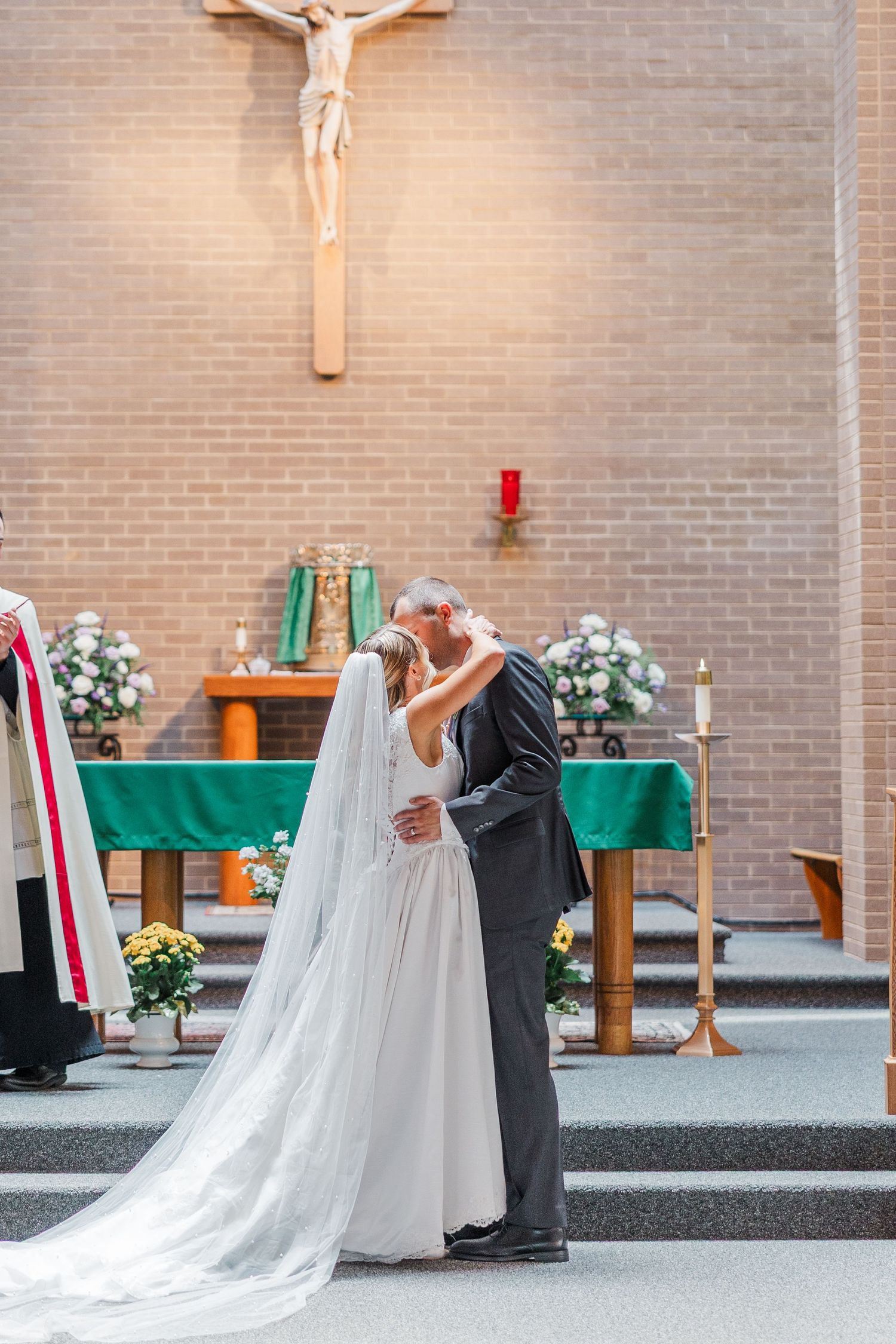 Bride and groom share first kiss at church altar with crucifix backdrop during Catholic wedding ceremony.