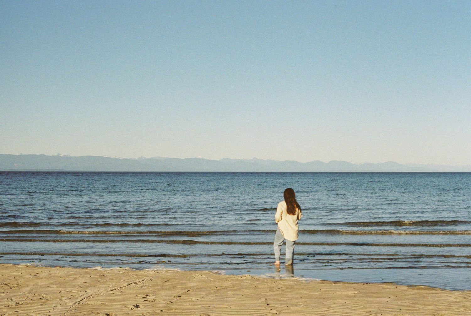 A woman in white stands at the shoreline of a calm beach, gazing at the serene blue ocean under a clear sky.