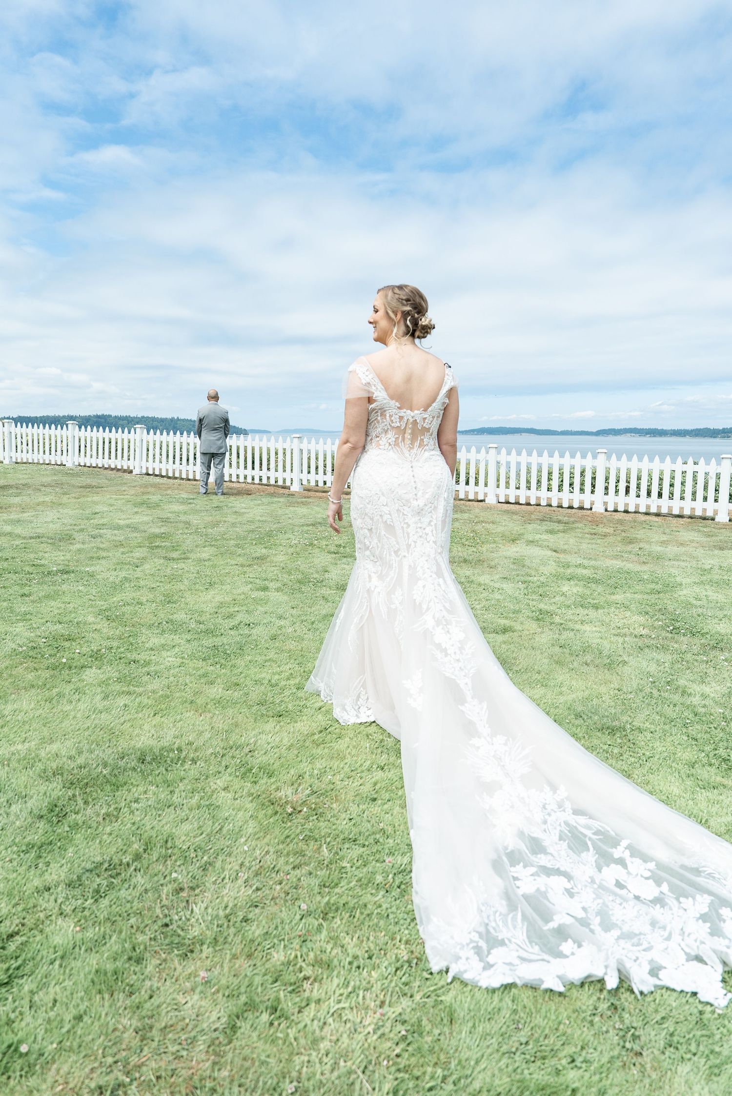 Bride in lace mermaid wedding gown with train stands on green lawn overlooking ocean with white picket fence.