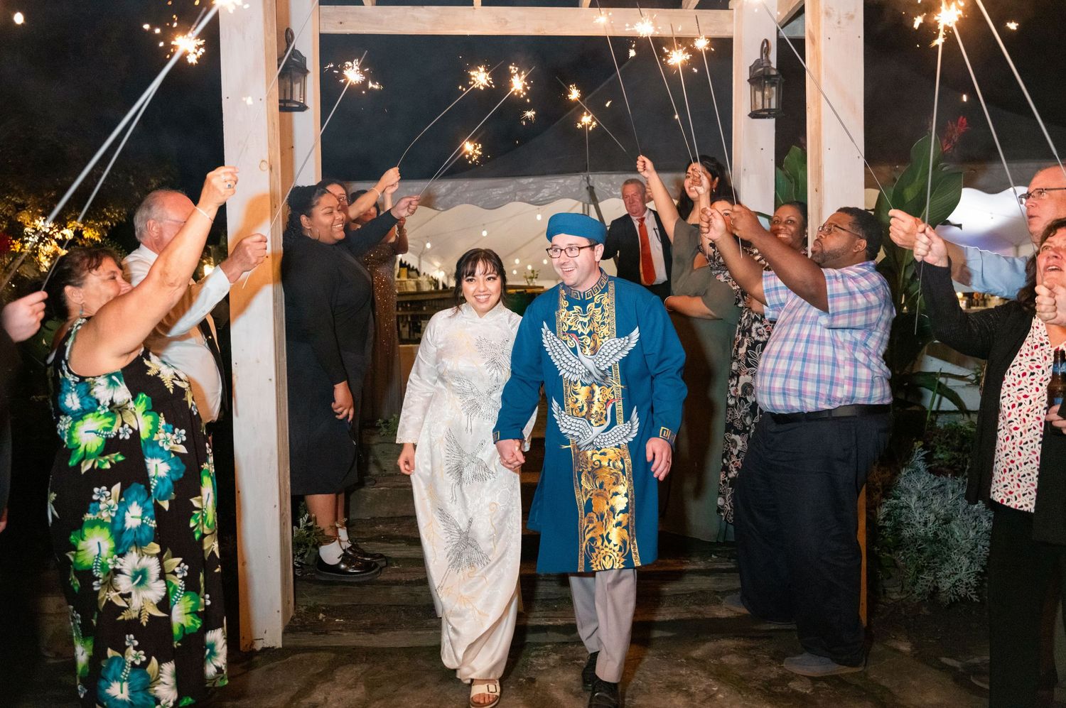 Jubilant couple walks under a sparkler send-off at nighttime wedding celebration with guests holding lit sparklers.