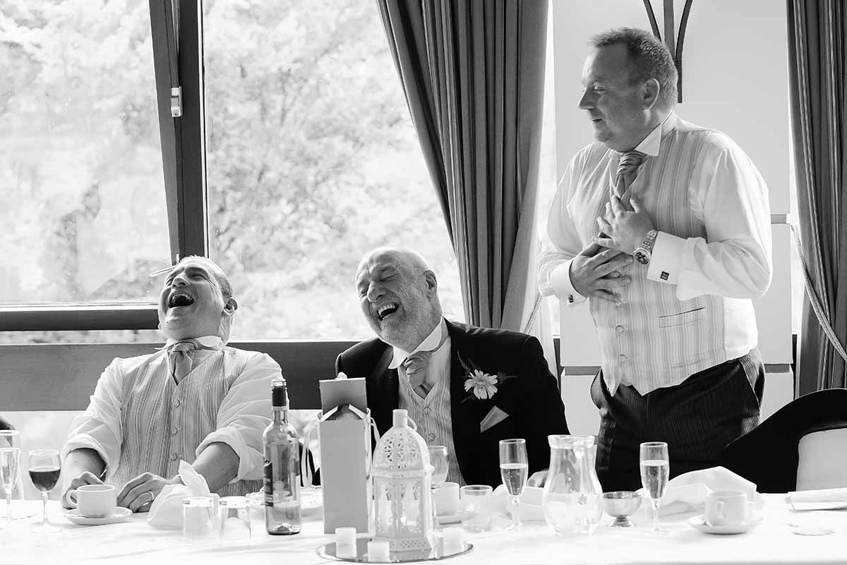 Black and white photo of three men laughing joyfully at a wedding reception dinner table.