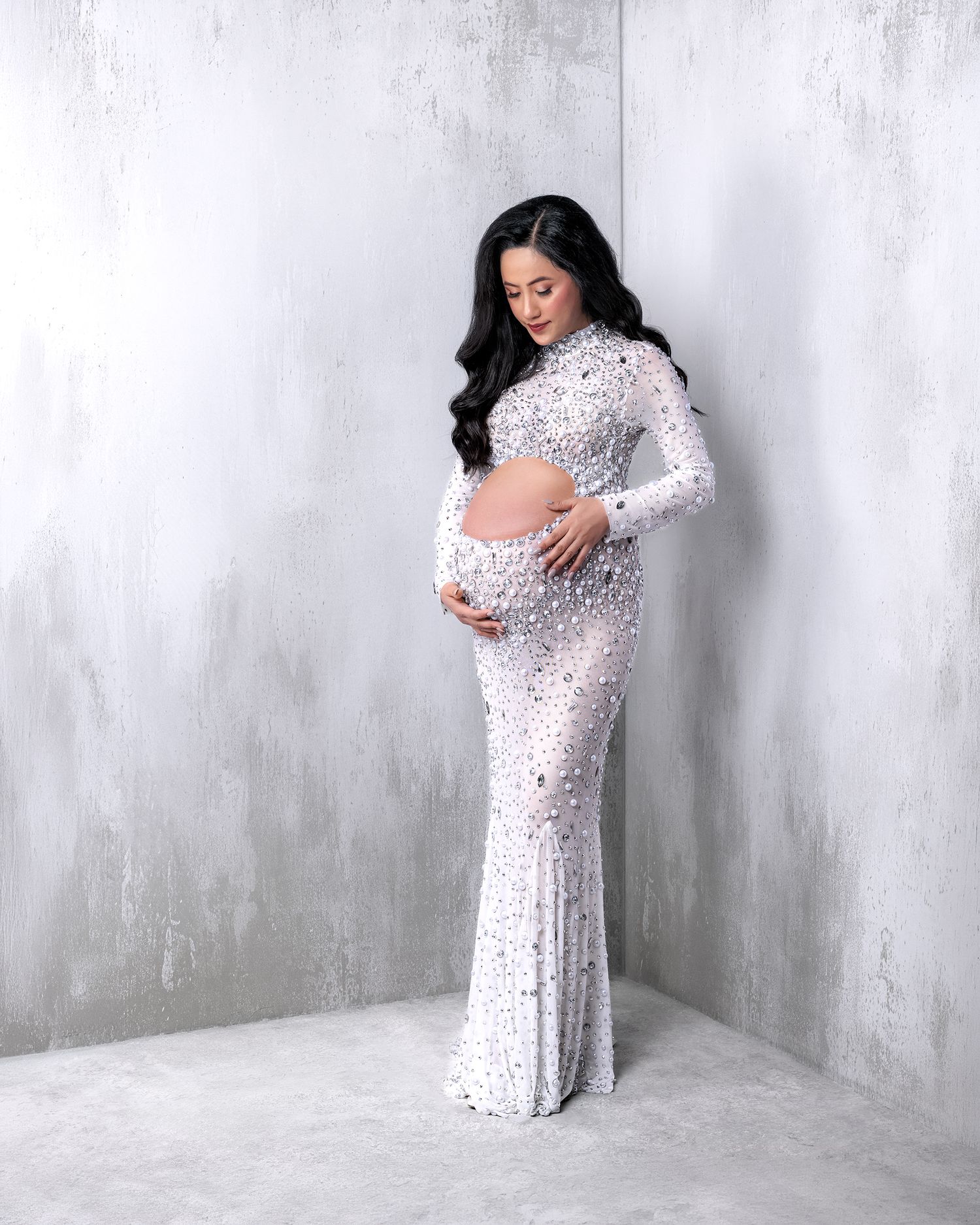 Maternity portrait of a beautiful woman in a figure hugging jewel encrusted gown against a modern white washed corner set backdrop.