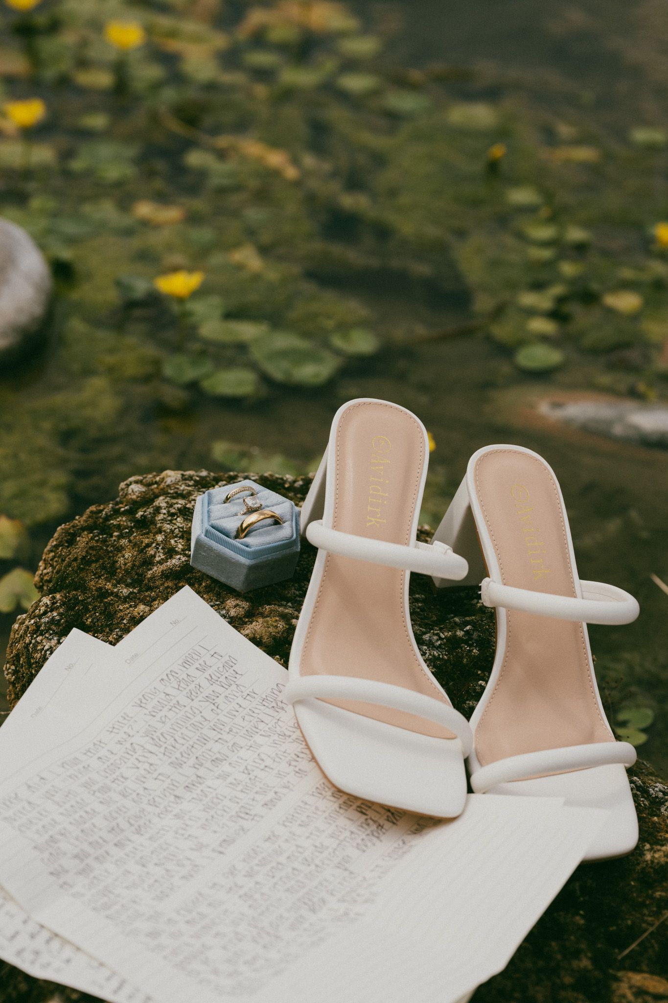 White bridal heels beside a velvet ring box with gold wedding bands and vows on a mossy rock near water.