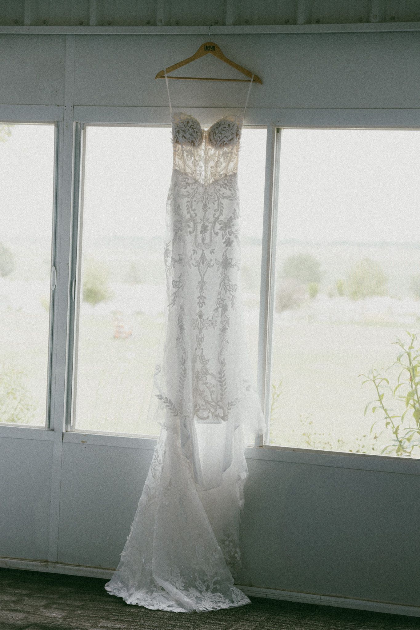 Elegant beaded lace wedding gown hanging on wooden hanger in sunlit window.