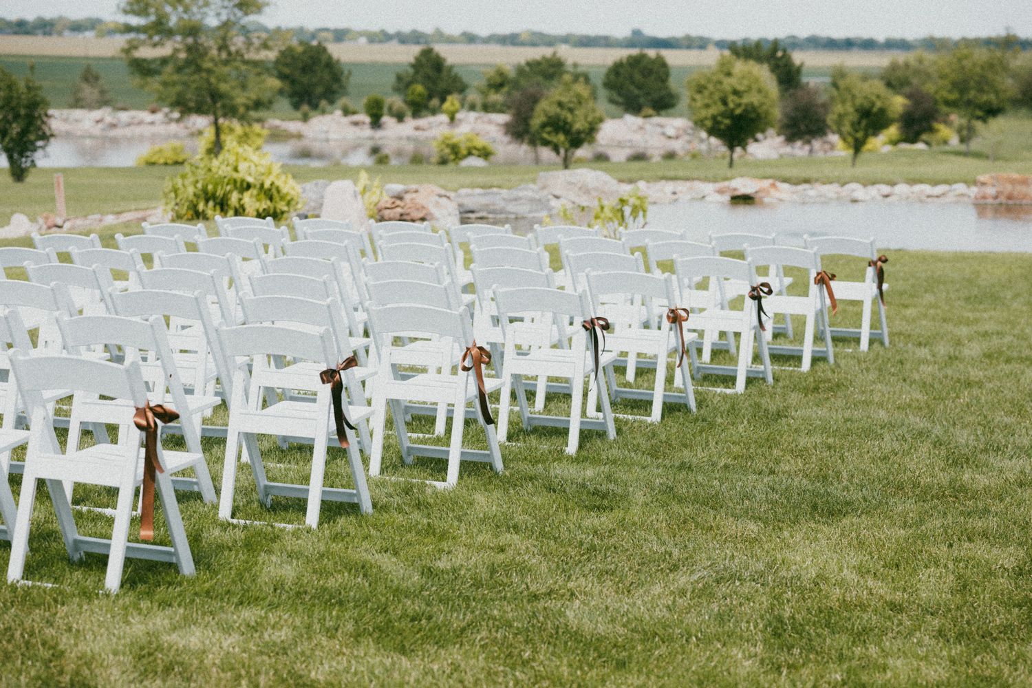 White folding chairs arranged in rows on green grass for an outdoor wedding ceremony with a scenic lake backdrop.