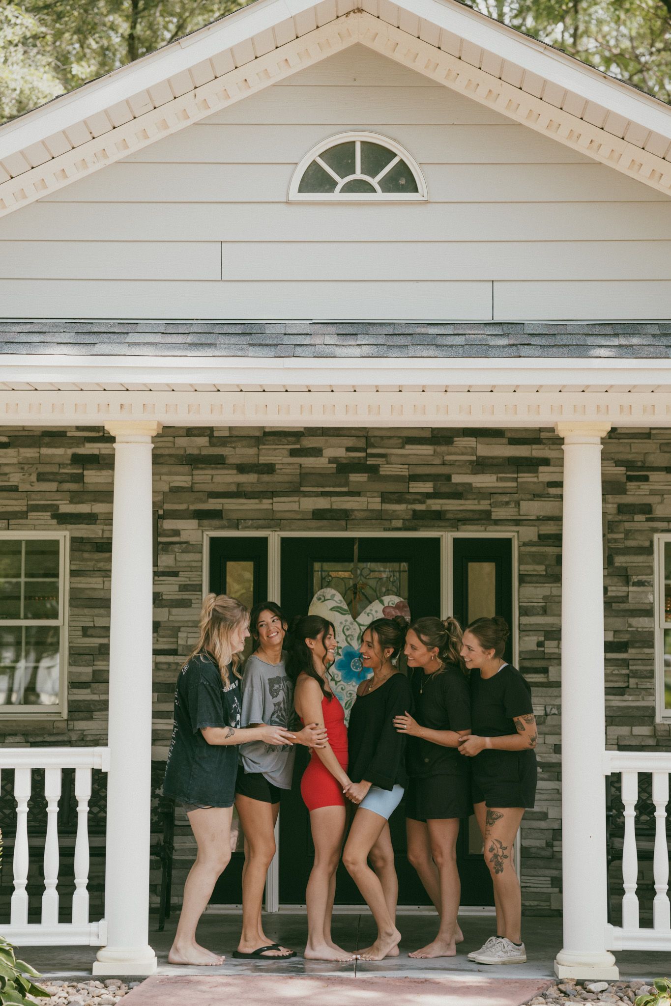 Group of women gathered on a charming brick and white house porch, socializing outdoors on a sunny day.