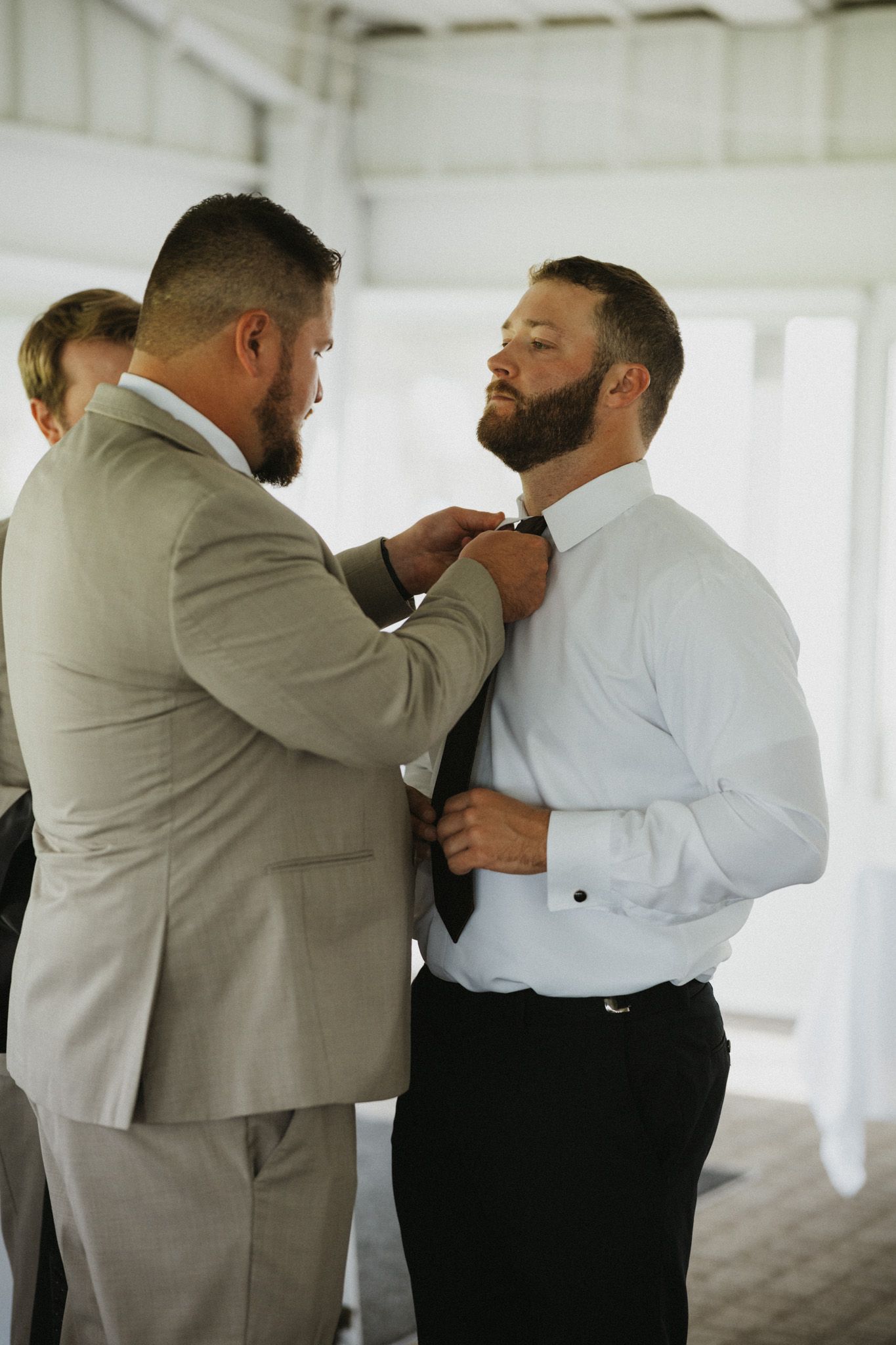 A groomsman in a tan suit adjusts the tie of a bearded groom in a white dress shirt before a wedding ceremony.