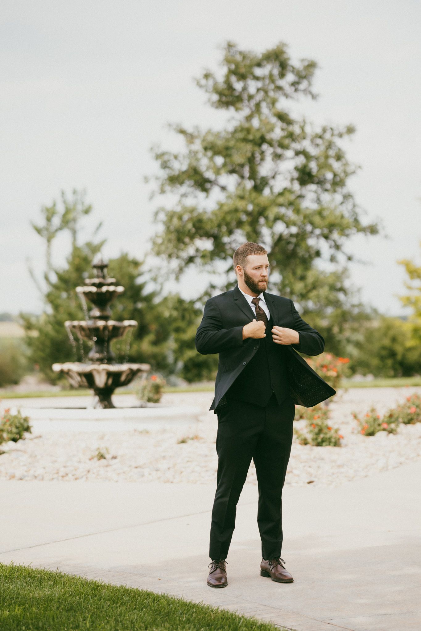 A bearded groom in a black tuxedo buttons his jacket while standing on a garden path with a fountain backdrop.