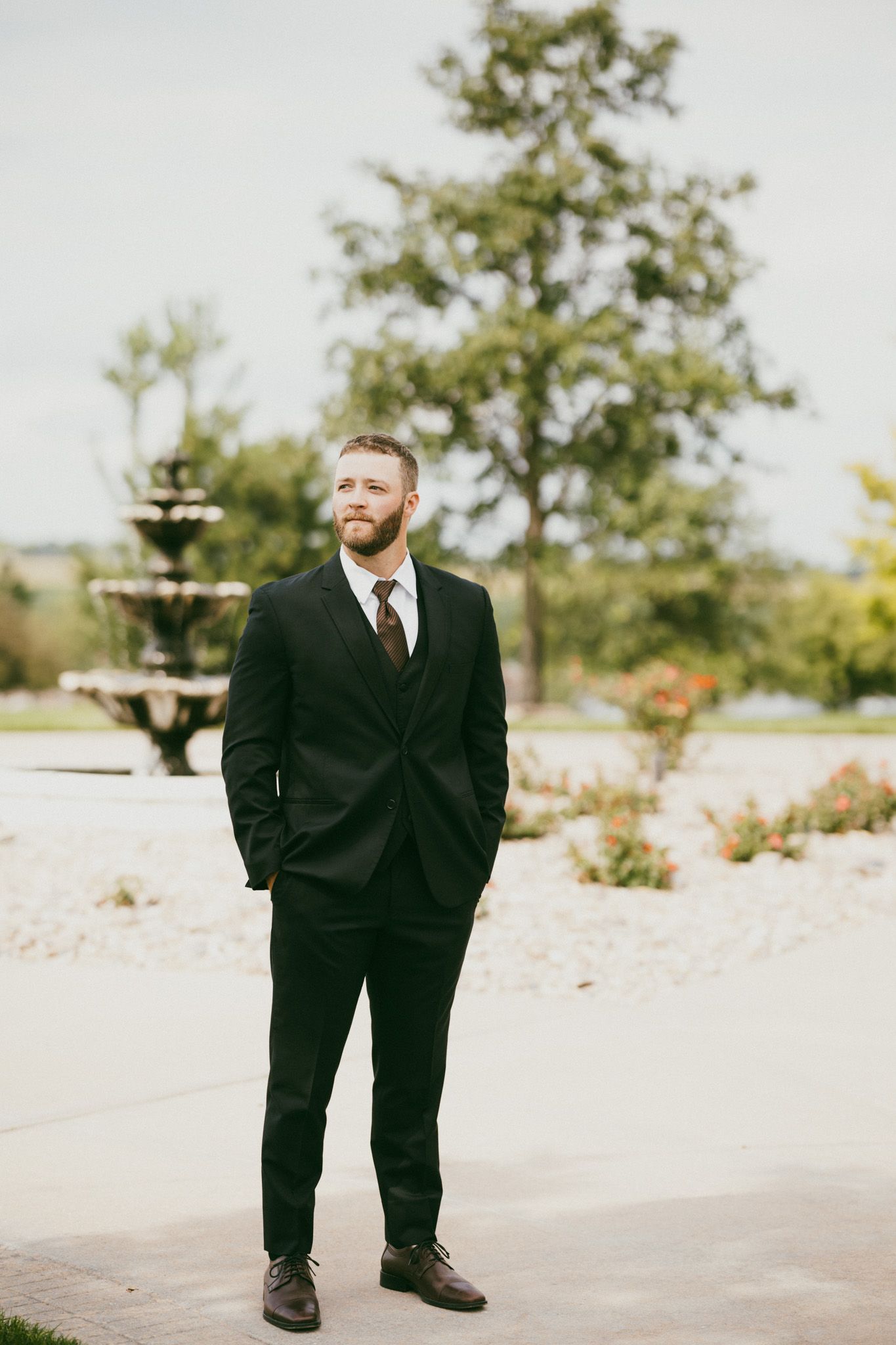 Bearded groom in black suit and tie stands confidently outdoors near a fountain and lush greenery.