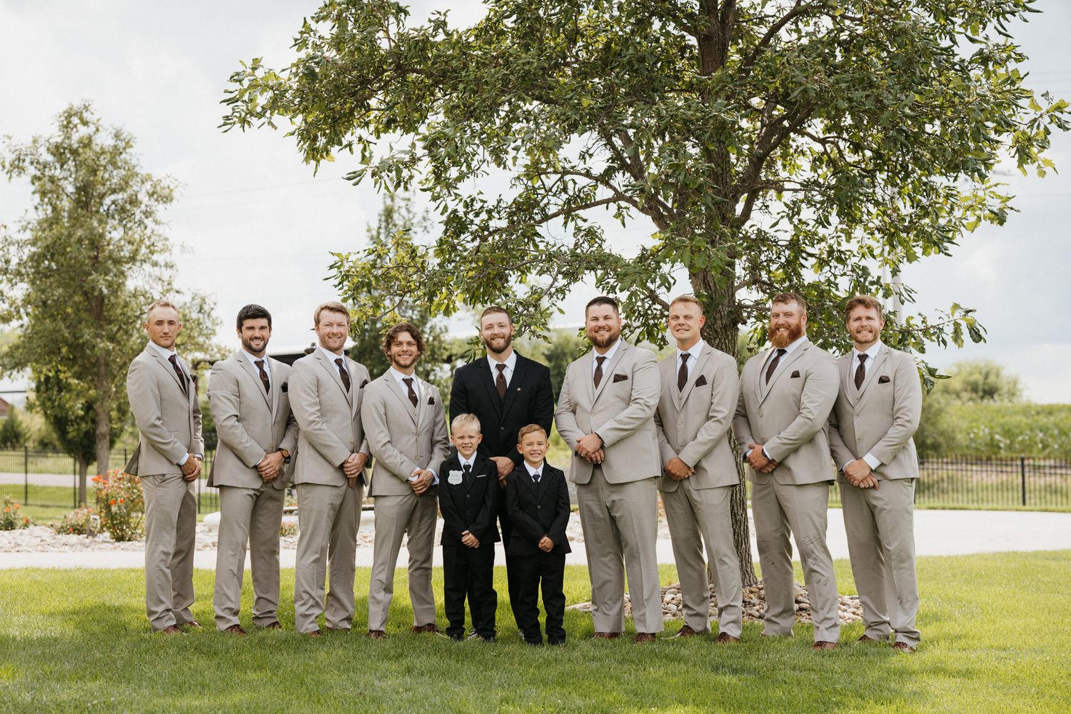 Groomsmen and groom posing in tan suits with black ties outdoors under a tree on a wedding day.