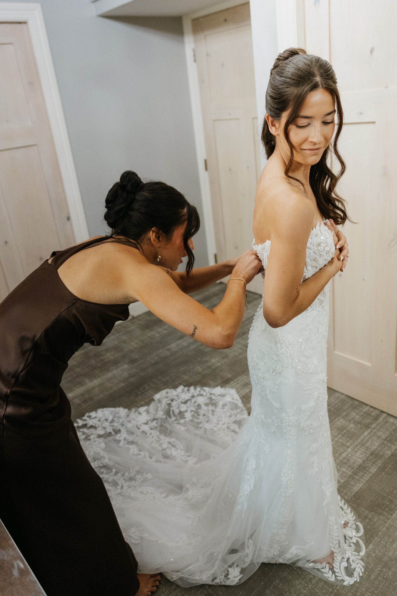 Bridesmaid helping bride button up her lace wedding dress before the ceremony.