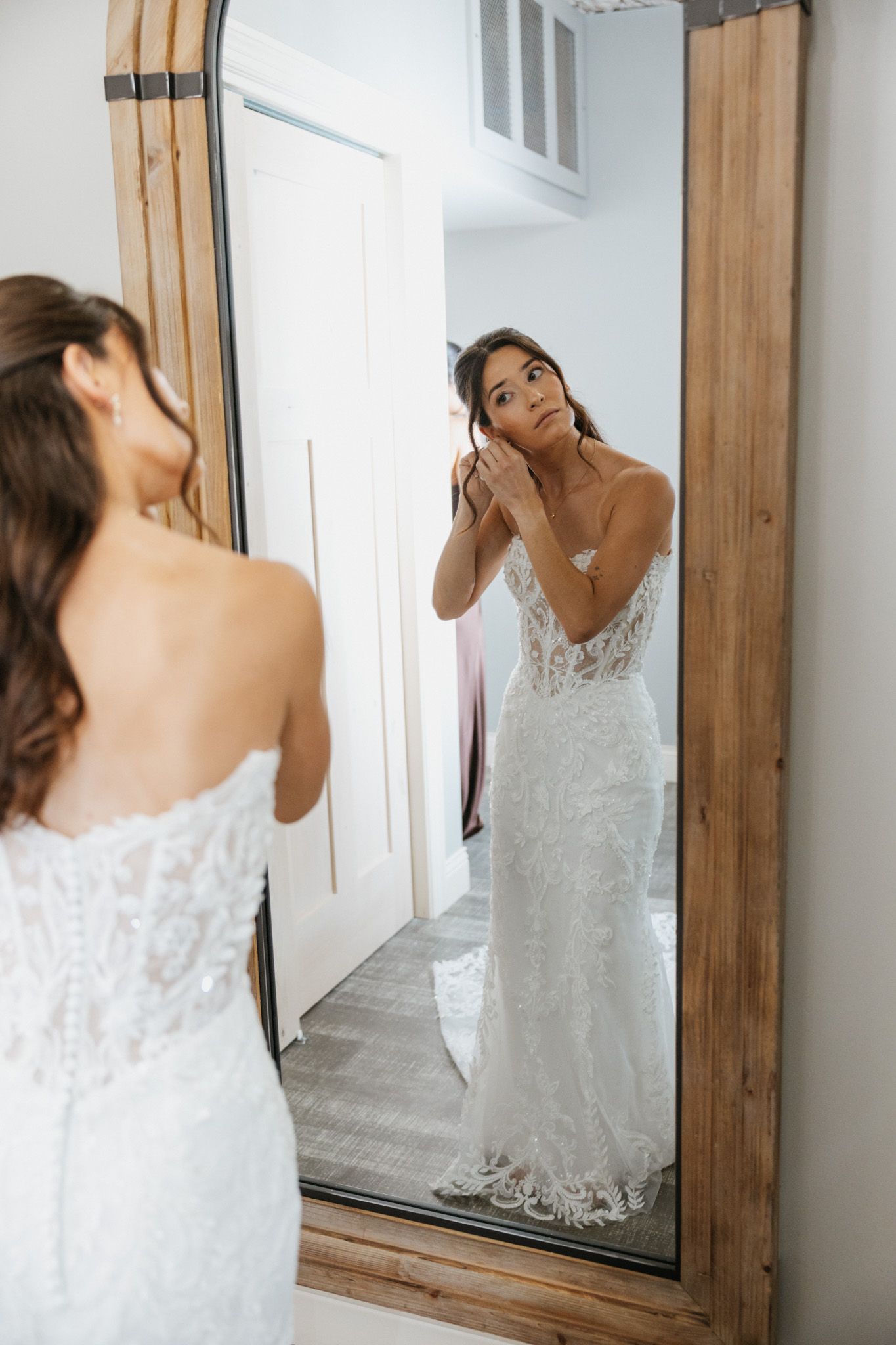 Bride in lace wedding gown adjusts earring while admiring her reflection in a large wooden mirror.