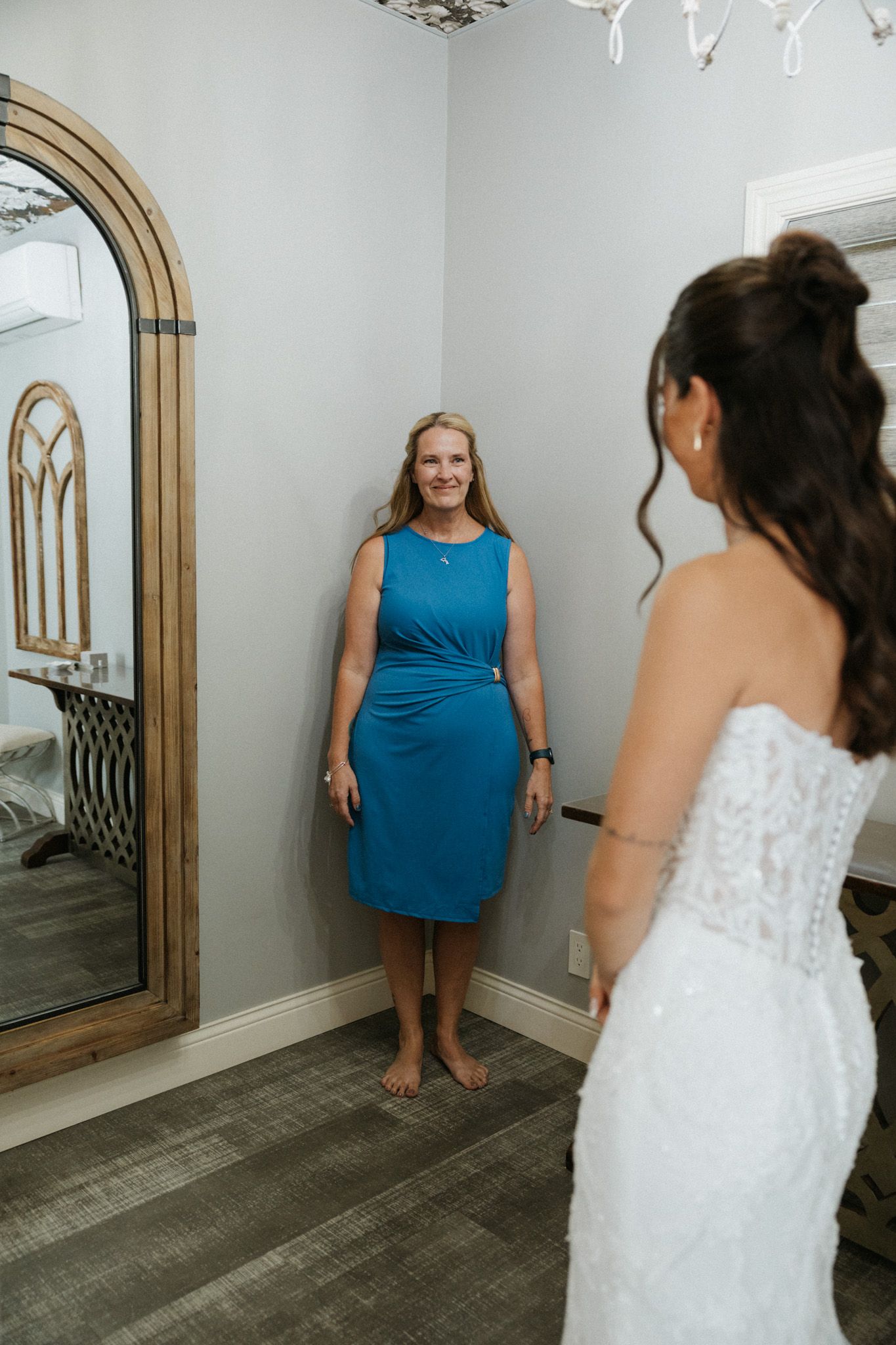 Bride in white lace gown shares an emotional first look moment with a woman in a blue dress in a bridal suite.