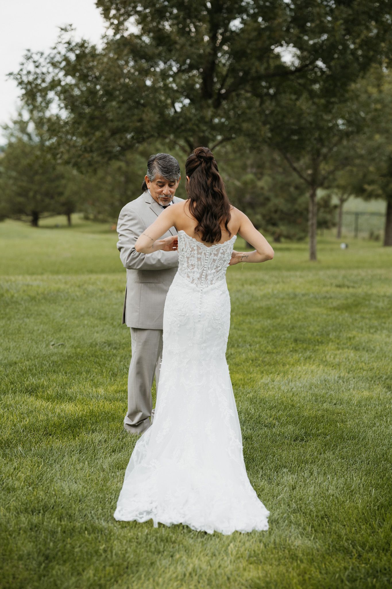 Groom in gray suit fastens the back of bride's white lace wedding gown outdoors on lush green grass.