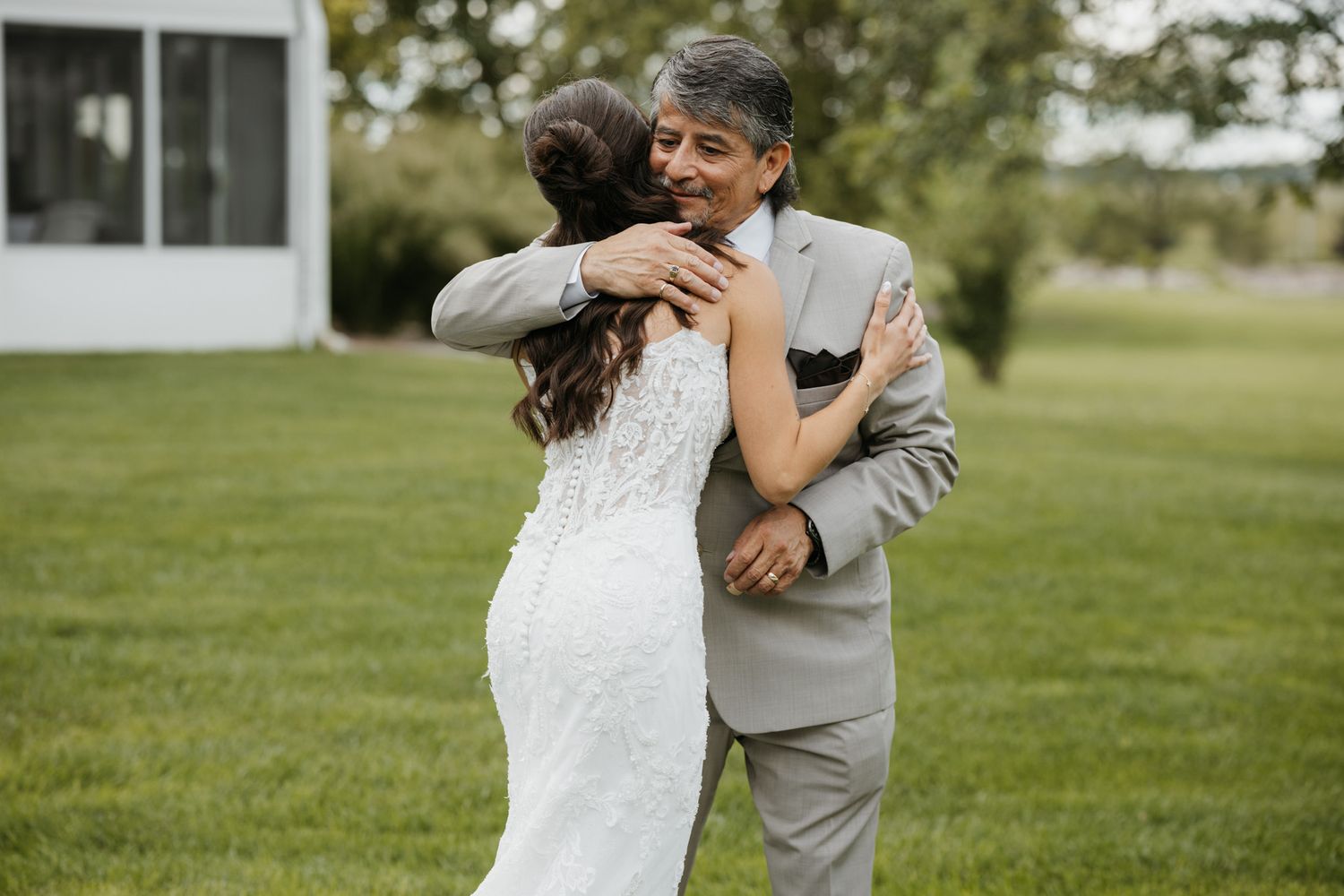 Bride in lace wedding dress sharing emotional hug with father on green lawn.