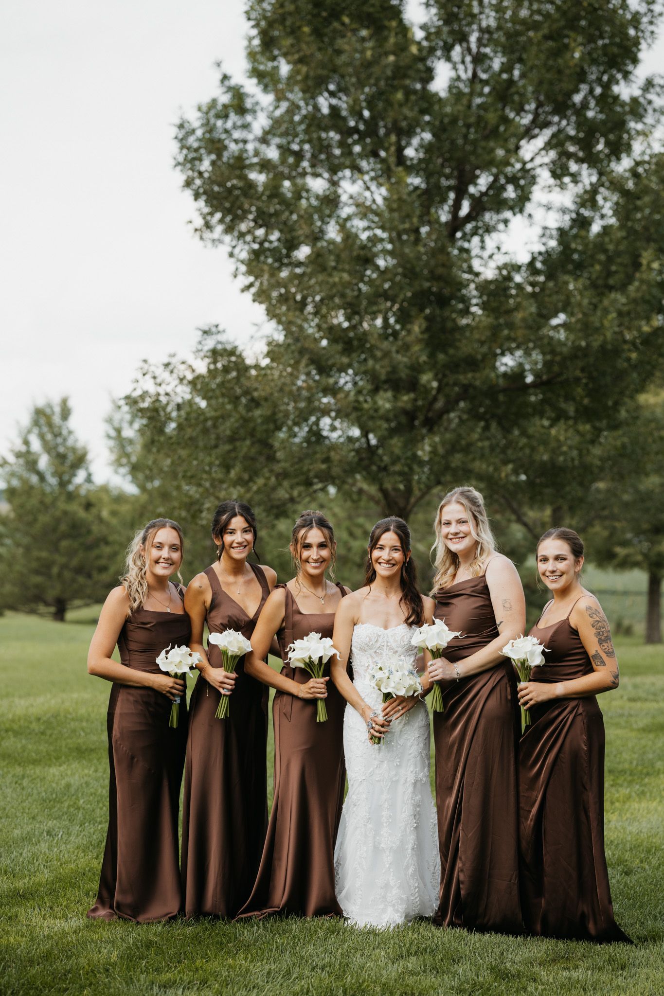 Bride in white lace gown poses with five bridesmaids in brown satin dresses holding white bouquets outdoors.