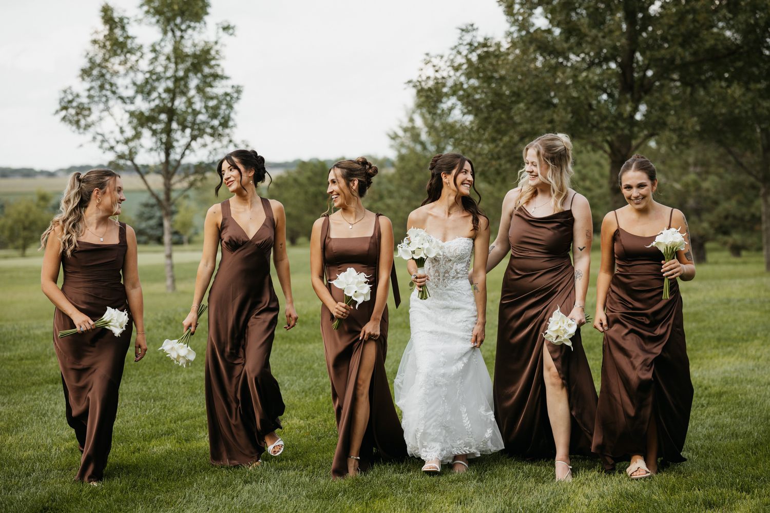 Bride walks with five bridesmaids wearing matching chocolate brown satin gowns across a green lawn.