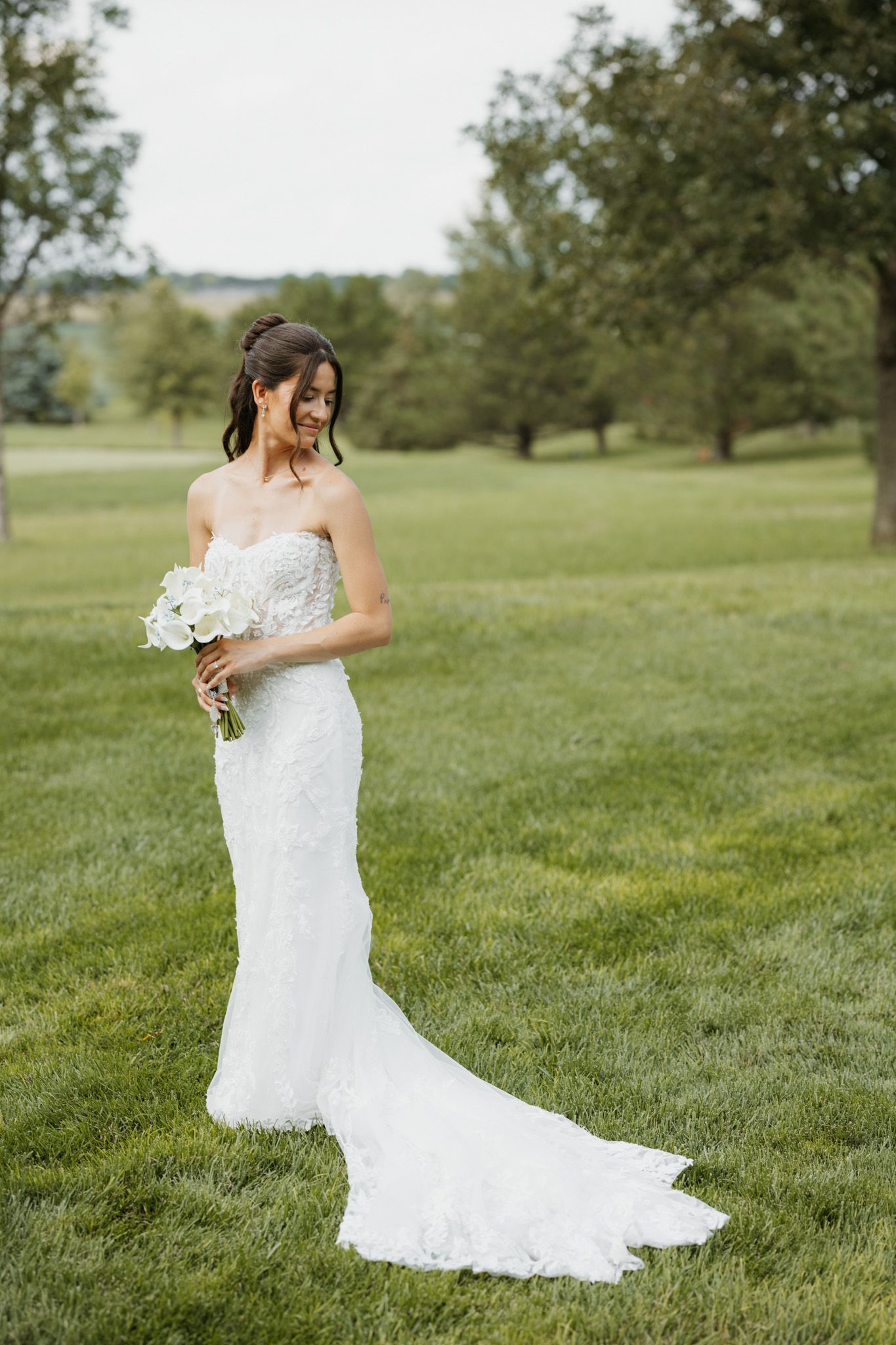 Elegant bride in strapless white lace mermaid gown holds white bouquet while standing on green grass.