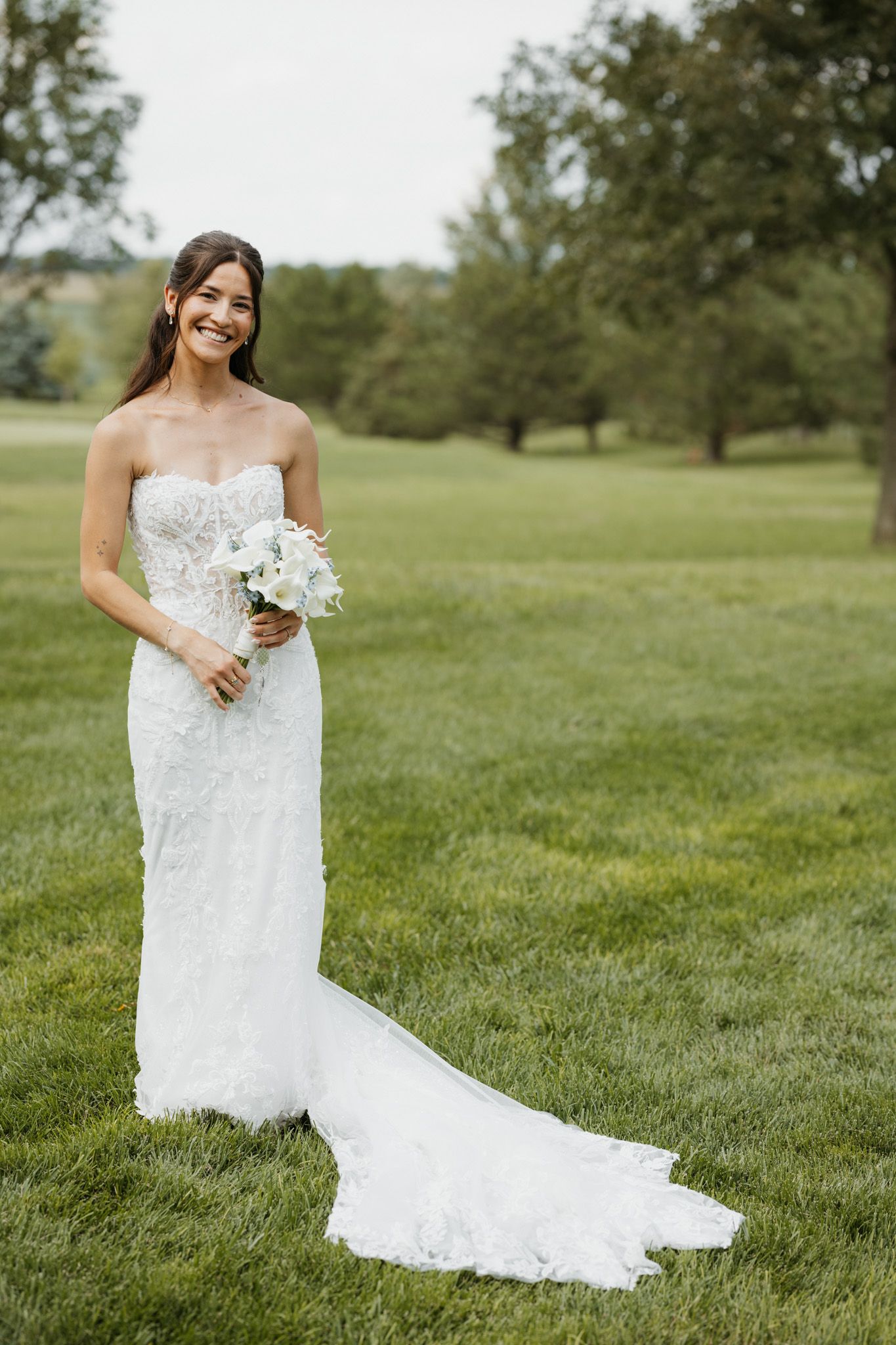 Smiling bride in strapless lace wedding gown holding white bouquet standing on green lawn outdoors.