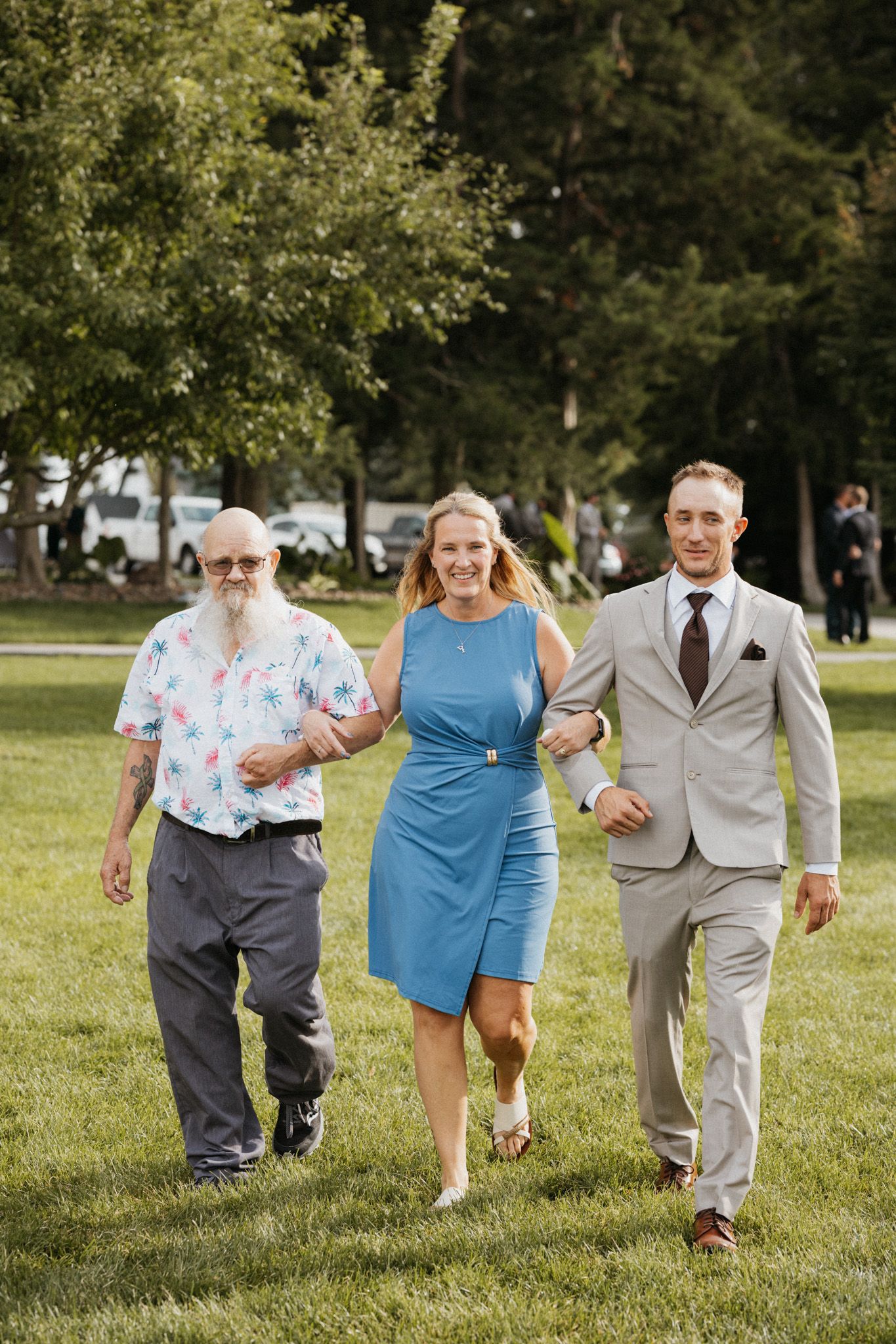 Three people walking on grass at outdoor wedding, woman in blue dress escorted by older man and groom.