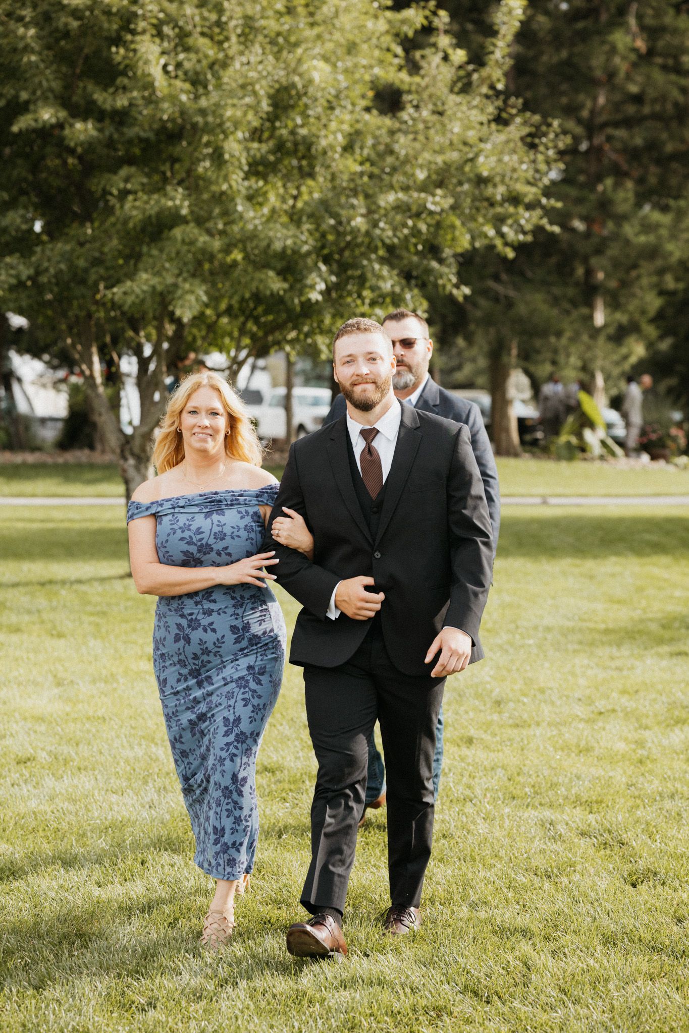 Groom escorted by his mother in a blue dress walking across green lawn at outdoor wedding.