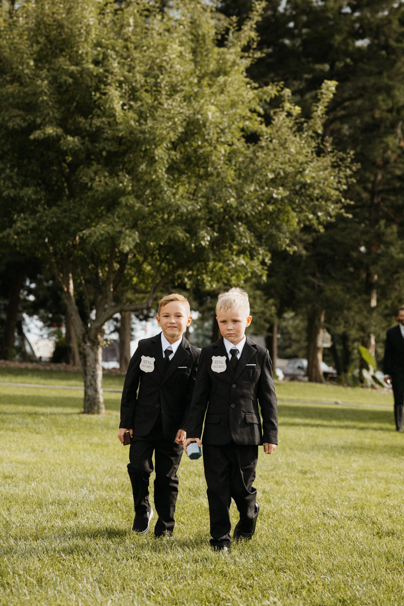 Two young ring bearers in matching black suits walking down the aisle at an outdoor wedding.