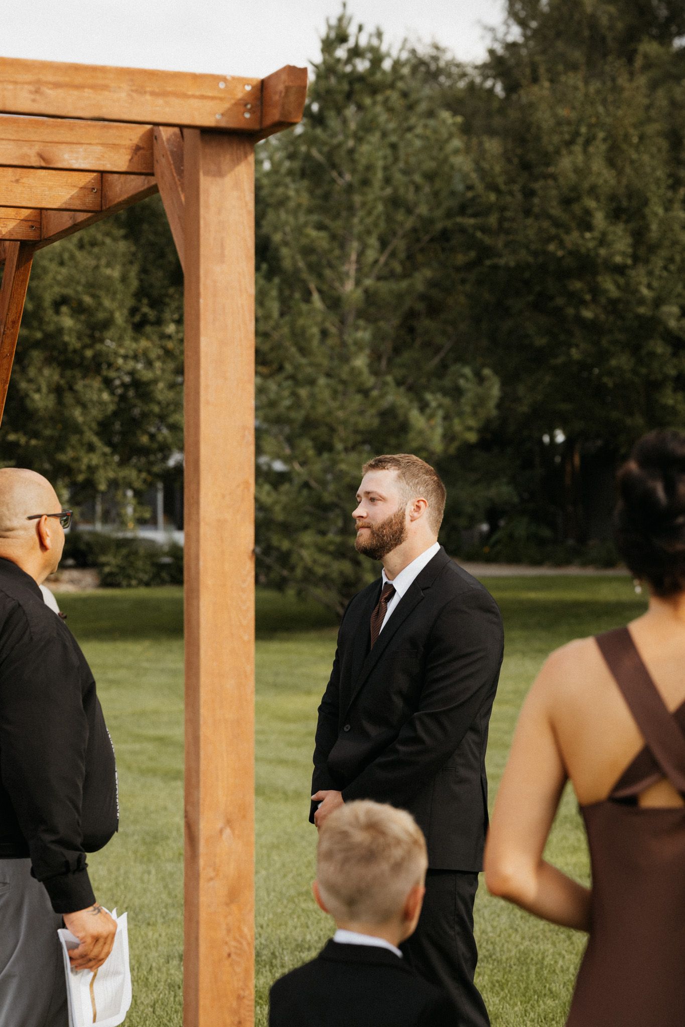 Emotional groom in black suit stands at outdoor wooden arch altar awaiting his bride.