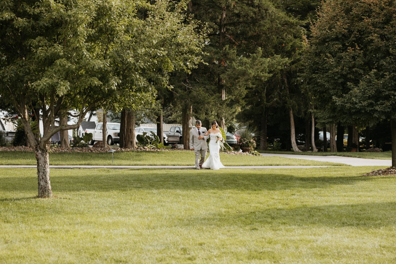 Bride in white gown walks with escort across lush green lawn toward outdoor wedding ceremony.