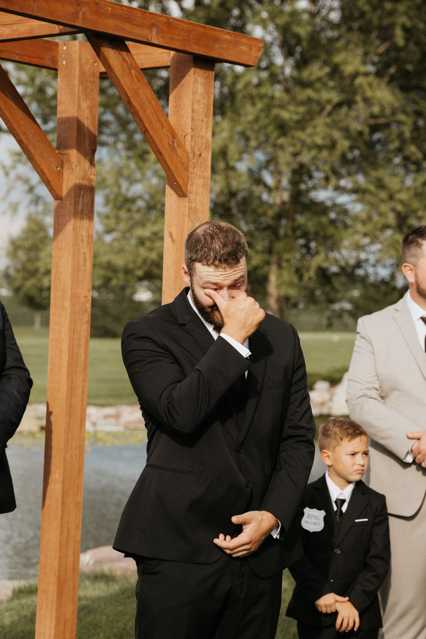 Tearful groom in black suit wipes eyes at outdoor wedding altar with young ring bearer nearby.