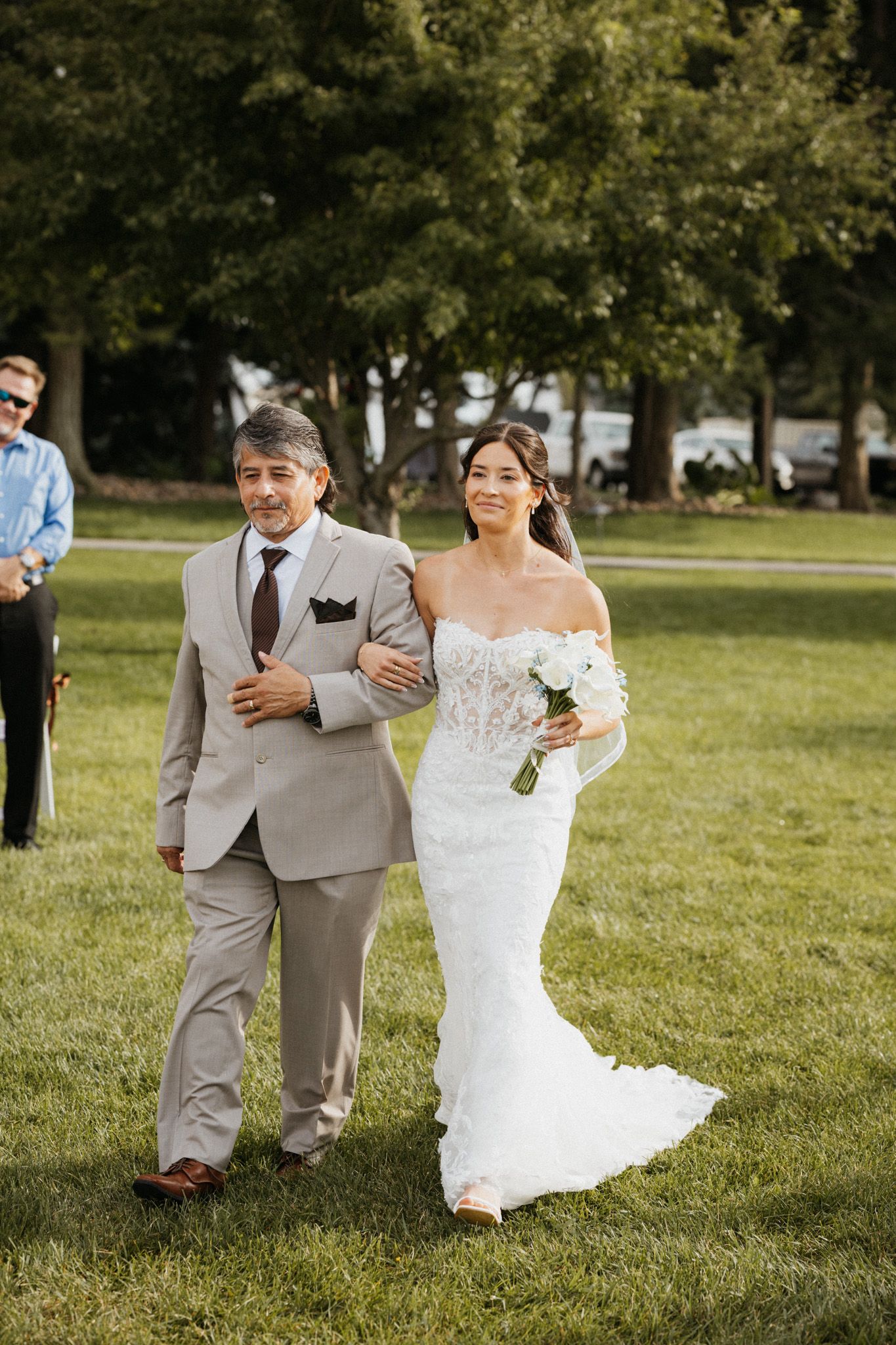 Bride in strapless lace gown walking down aisle on grass escorted by man in tan suit at outdoor wedding.