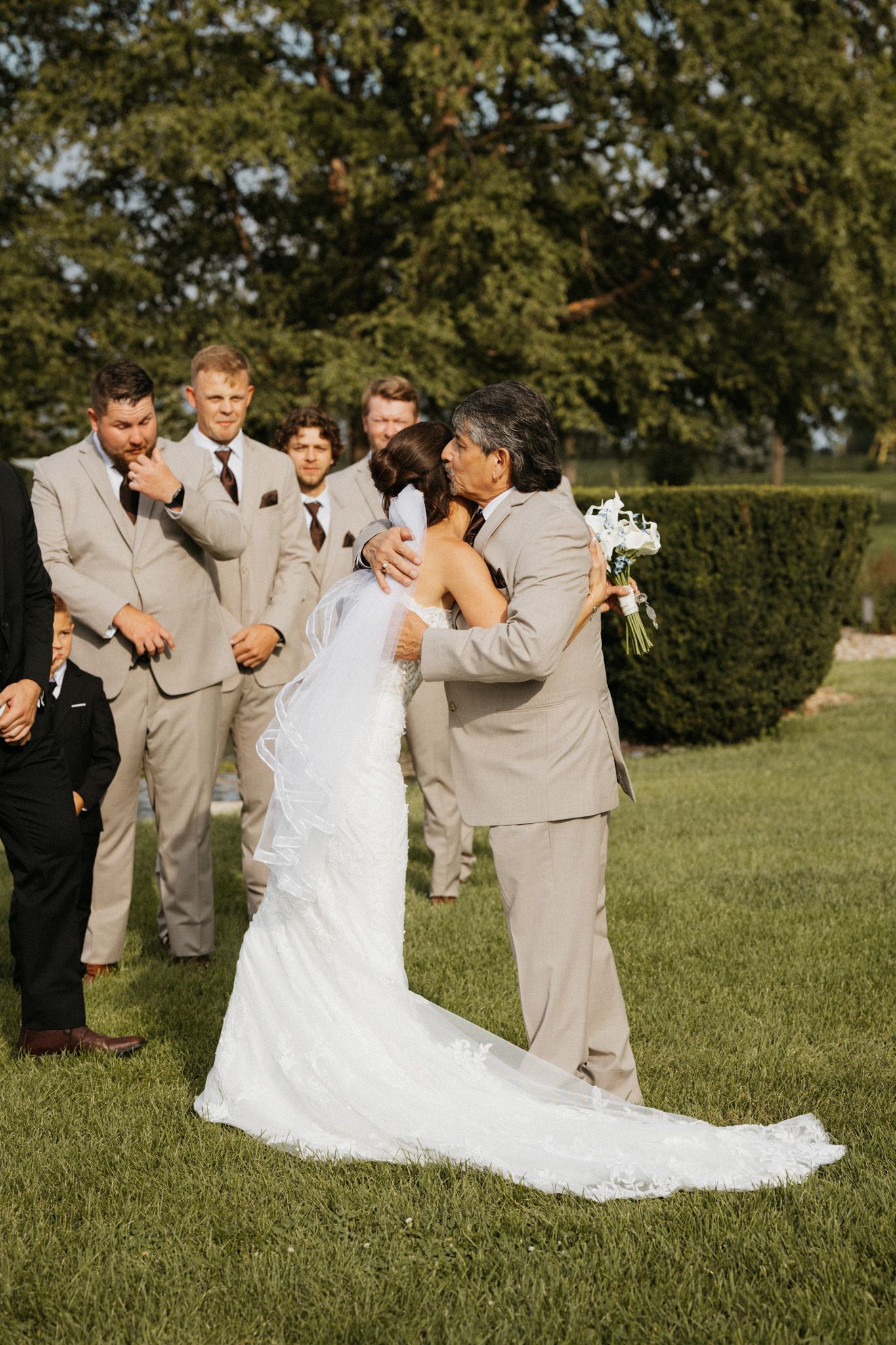 Father embraces bride in white lace gown at outdoor ceremony as groomsmen in tan suits look on.