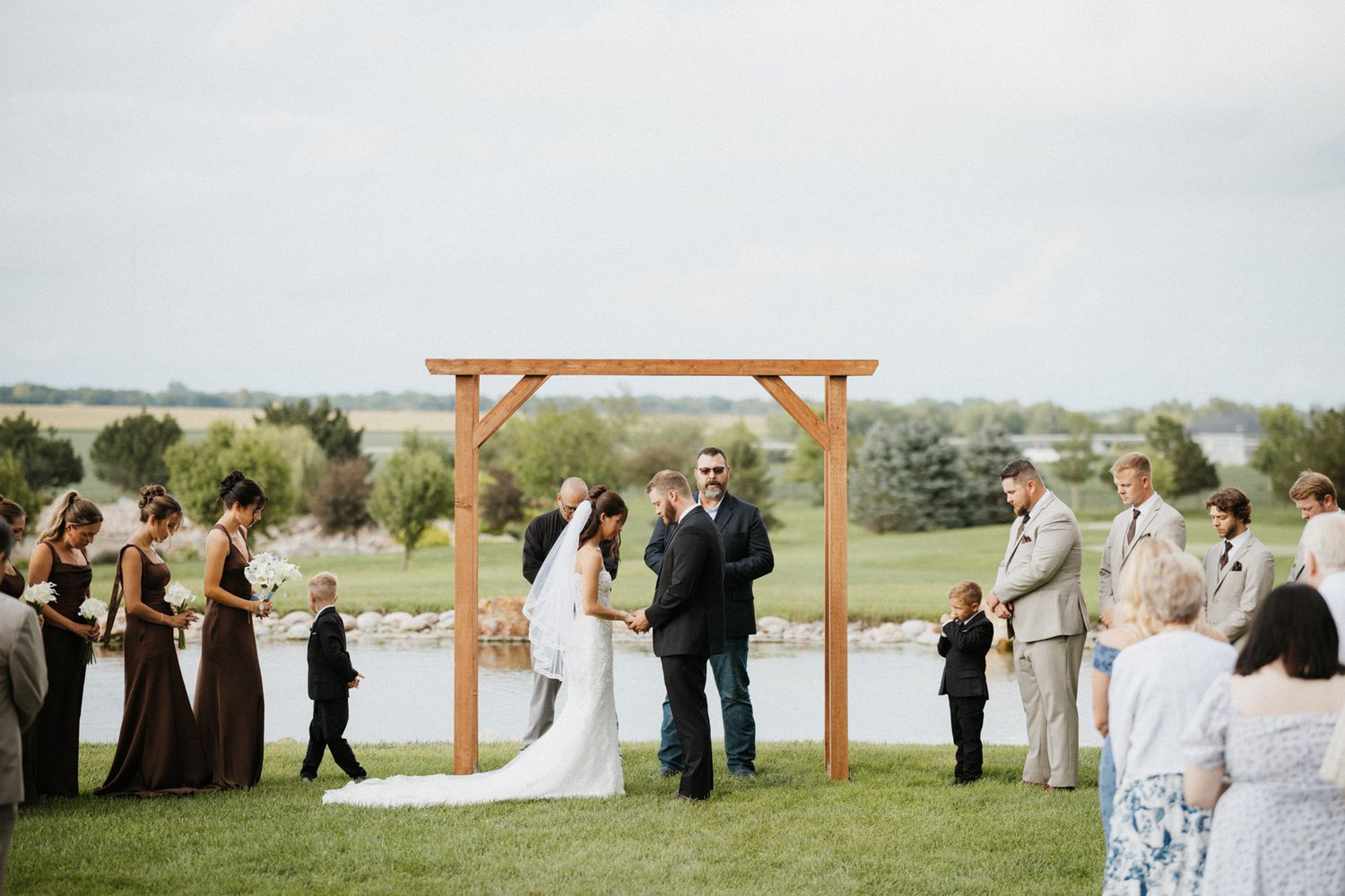 Bride and groom share a kiss under a wooden arch during an outdoor lakeside wedding ceremony.