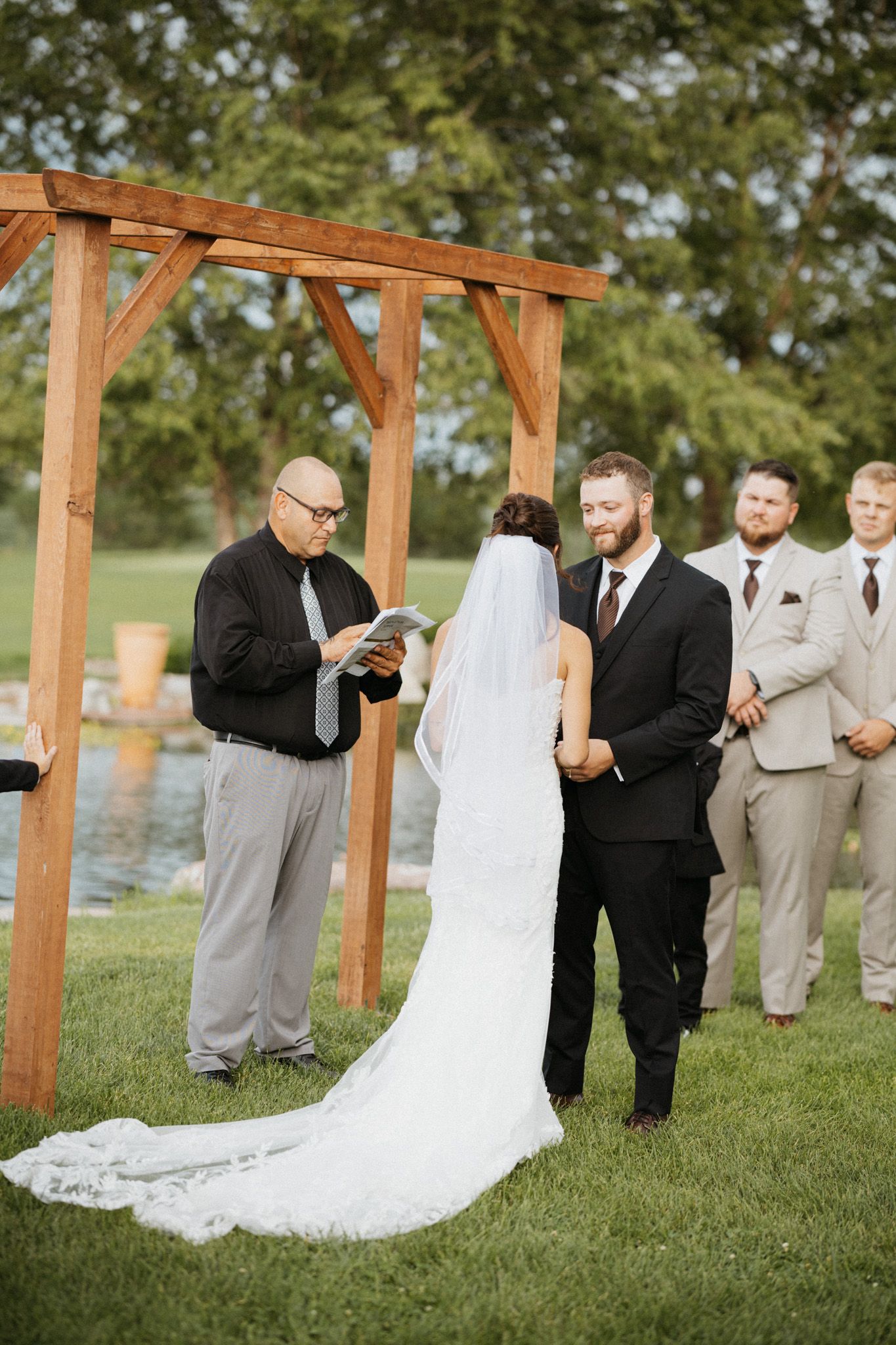 Outdoor wedding ceremony with couple exchanging vows under wooden arch near pond with groomsmen in tan suits.