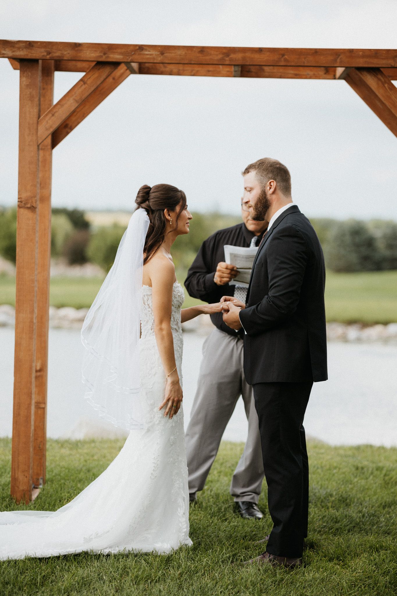 Bride and bearded groom exchanging vows under rustic wooden arch at outdoor waterfront wedding ceremony.