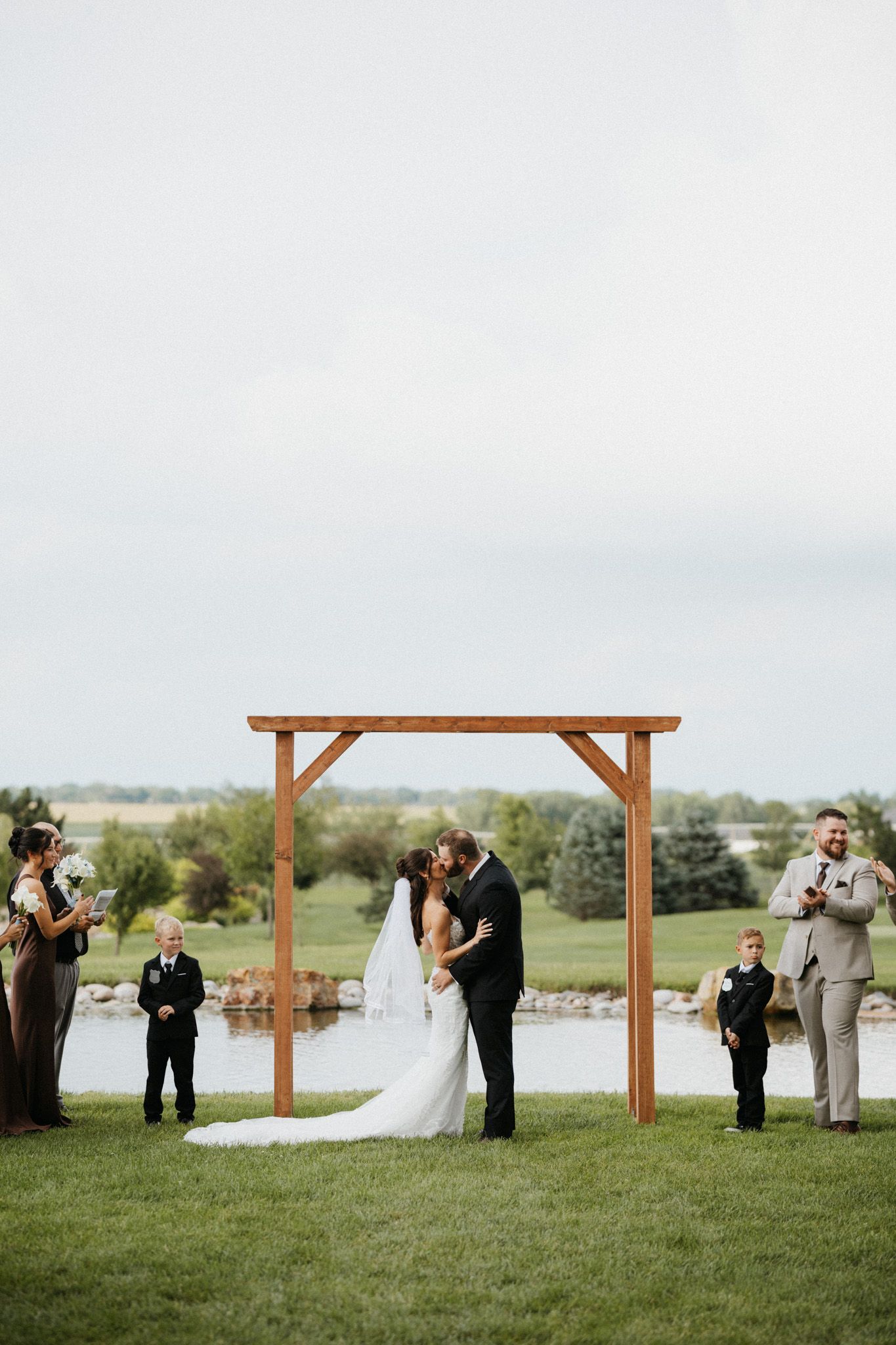Bride and groom sharing first kiss under wooden arch at outdoor wedding ceremony beside a scenic pond.