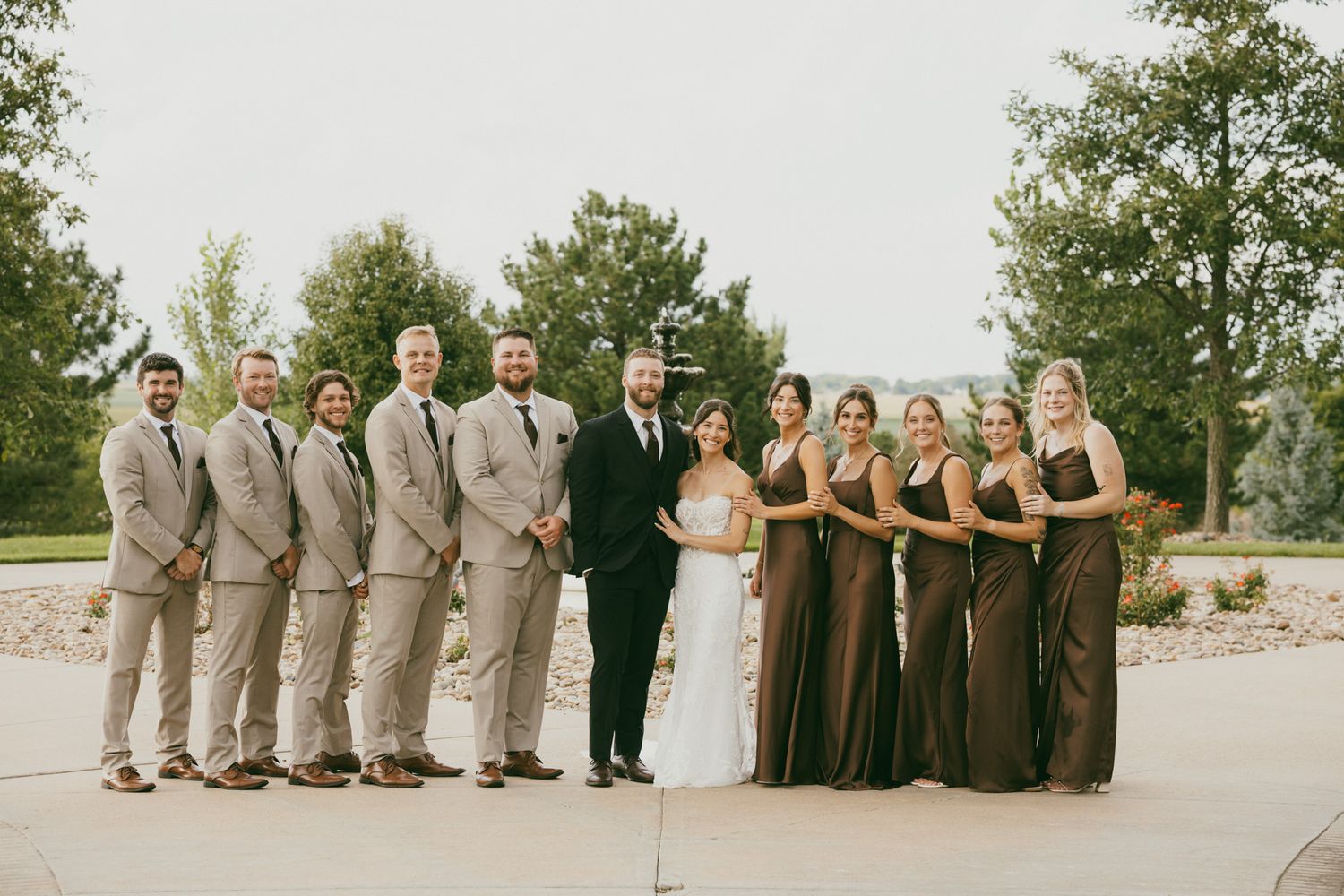 Wedding party poses together outdoors with groomsmen in tan suits and bridesmaids in brown satin gowns.